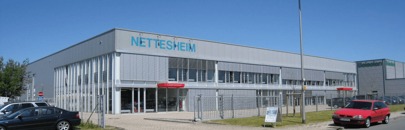 nettesheim Firmengebäude Modernes Firmengebäude der Firma Nettesheim mit großem Logo auf der Fassade. Glasfront mit Eingangsbereich, darüber ein Schild mit der Aufschrift ‚Lagerverkauf‘. Auf dem Parkplatz davor stehen mehrere Autos.