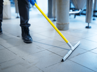 Eine Person mit schwarzen Arbeitsschuhen und blauen Handschuhen schiebt mit einem Bodenabzieher Wasser oder Reinigungsflüssigkeit über einen gefliesten Industrieboden. Im Hintergrund stehen große Edelstahlanlagen.