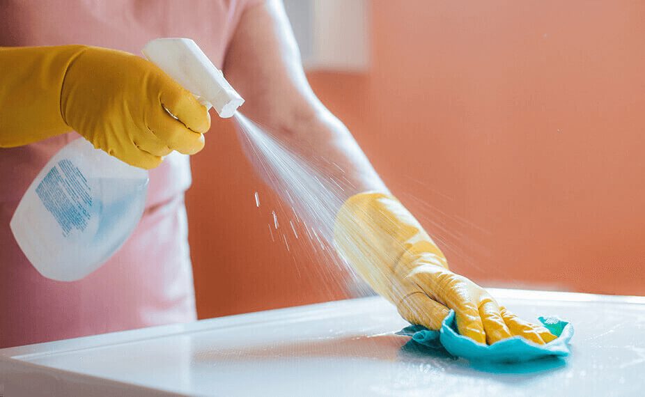 Person mit gelben Gummihandschuhen sprüht Reinigungsmittel auf eine glatte Oberfläche und wischt gleichzeitig mit einem blauen Tuch