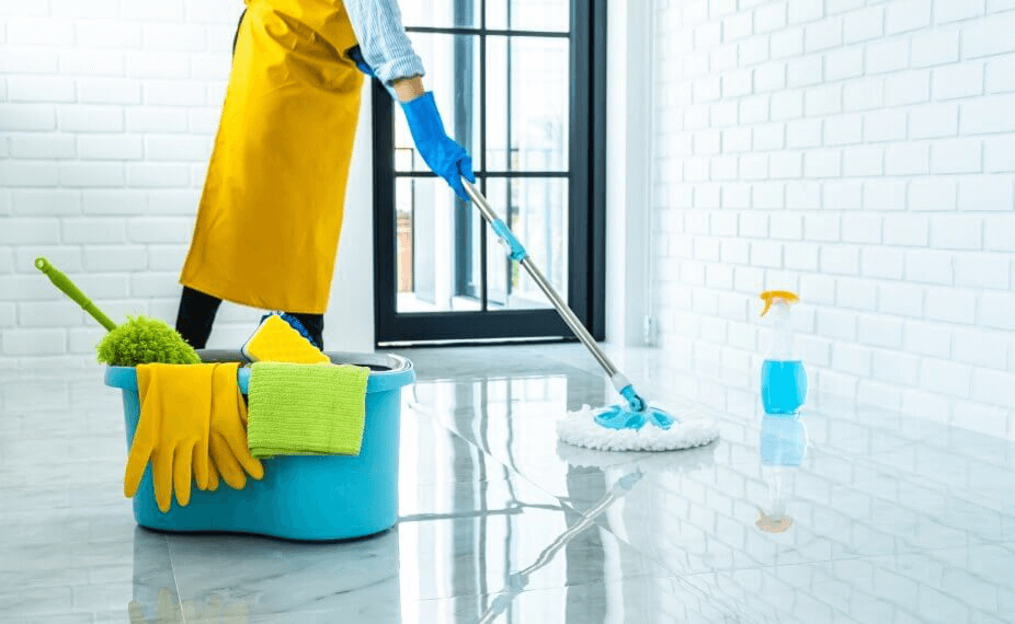 Person mit gelber Schürze und blauen Handschuhen wischt einen glänzenden Fliesenboden mit einem Flachmopp. Daneben stehen ein blauer Eimer mit Reinigungstüchern, Gummihandschuhen und Reinigungsutensilien sowie eine Sprühflasche mit blauer Flüssigkeit.