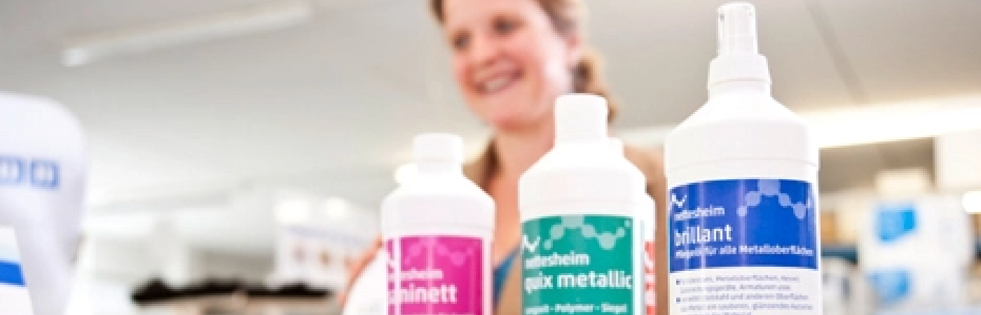 Kontaktaufnahme nettesheim Drei Reinigungsmittel-Flaschen von Nettesheim mit farbigen Etiketten in Pink, Grün und Blau stehen im Vordergrund. Im Hintergrund lächelt eine unscharf dargestellte Frau in einem hellen Verkaufsraum.